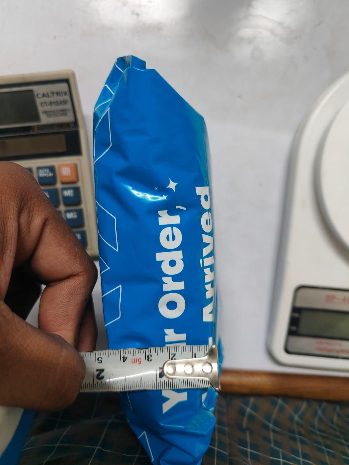 Blue package with 'Your order has arrived' text, held by a hand with a ruler for scale, on a white surface with a calculator and digital scale.