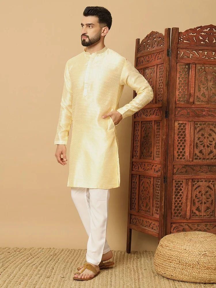 Man wearing a cream kurta with white pants standing next to a wooden carved cabinet.