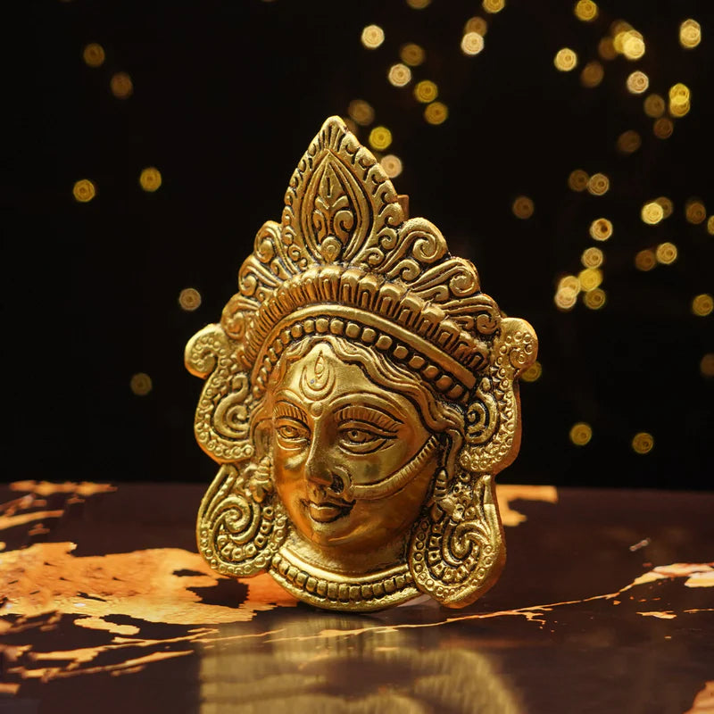 Decorative gold mask with intricate designs on a reflective surface with a dark background.